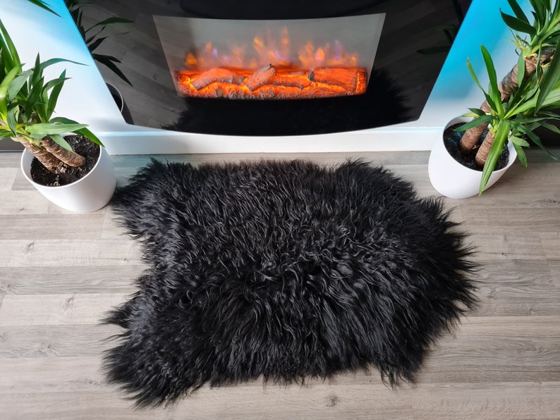 May include: A plush, long-haired black sheepskin rug with a thick, shaggy texture and an irregular, natural pelt shape. The rug features dense, wavy fibers that create a deep, dark appearance, providing a soft and voluminous surface covering.