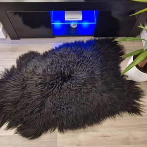 May include: A black sheepskin rug with long, thick, and curly fur. The rug has an irregular, organic shape with a dense texture and a deep, solid black color throughout the entire piece, showcasing the natural length and volume of the fibers.