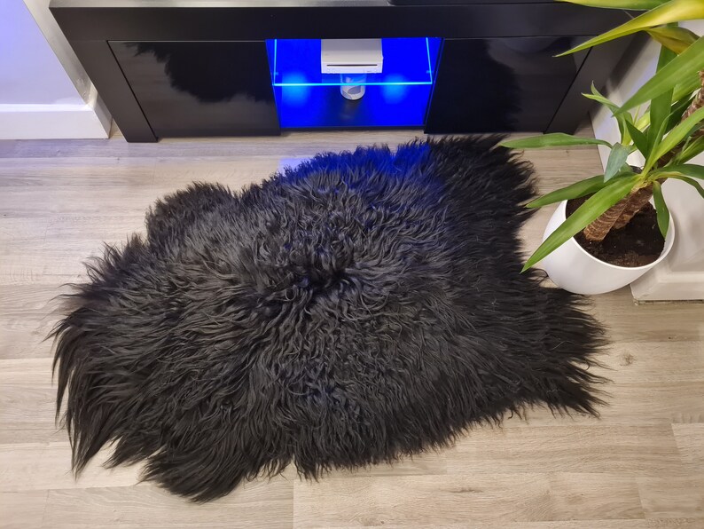 May include: A black sheepskin rug with long, thick, and curly fur. The rug has an irregular, organic shape with a dense texture and a deep, solid black color throughout the entire piece, showcasing the natural length and volume of the fibers.