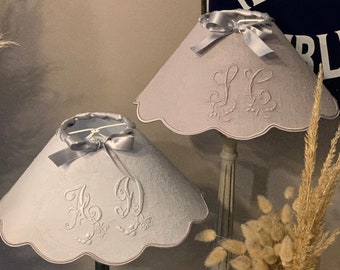 May include: Two gray lampshades with scalloped edges and embroidered initials. The lampshades are on black and gray lamp bases. The lampshades are decorated with silver satin bows. The background includes a sign that reads "Téléphone Public" and a vase with dried pampas grass.