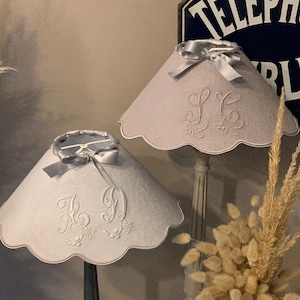 Könnte beinhalten: Zwei graue Lampenschirme mit gewellten Rändern und gestickten Initialen. Die Lampenschirme stehen auf schwarzen und grauen Lampenfüßen. Die Lampenschirme sind mit silbernen Satinbändern verziert. Der Hintergrund zeigt ein Schild mit der Aufschrift "Téléphone Public" und eine Vase mit getrocknetem Pampasgras.