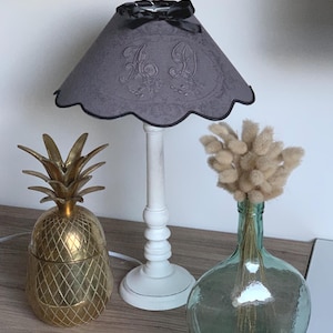 Könnte beinhalten: Eine dekorative Szene mit einem goldenen ananasförmigen Behälter, einem grauen Lampenschirm mit den Buchstaben "A D" und einer klaren Glasflasche mit getrockneten Blumen. Die Lampe hat einen weißen Sockel und eine schwarze Schleife.