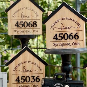 May include: Wooden house-shaped signs with black trim and the phrase "THERE'S NO PLACE LIKE home" above a number and city name. The signs display the numbers 45068, 45066, and 45036, with the cities Waynesville, Springboro, and Lebanon, Ohio. A black toy truck is also visible.