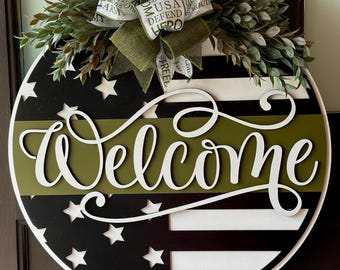 Peut inclure: Un rond de porte en bois avec un design de drapeau américain noir et blanc. Le drapeau a une bande verte au milieu avec le mot "Bienvenue" écrit en cursive blanche. Un nœud vert avec un ruban blanc est attaché au sommet du rond de porte.