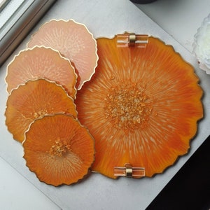 Peut inclure: Ensemble de quatre sous-verres en résine orange avec des accents dorés. Le plus grand sous-verre est un plateau avec deux poignées dorées.