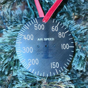 May include: A black, circular Christmas ornament resembling an airspeed indicator, with white numerals and text. The ornament is suspended by a red ribbon against a backdrop of a green Christmas tree.
