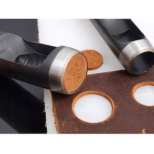 May include: Two black metal leather hole punch tools with a silver rim. The tools are on a brown leather surface with several round holes punched in it. There are also two brown leather circles on the surface.