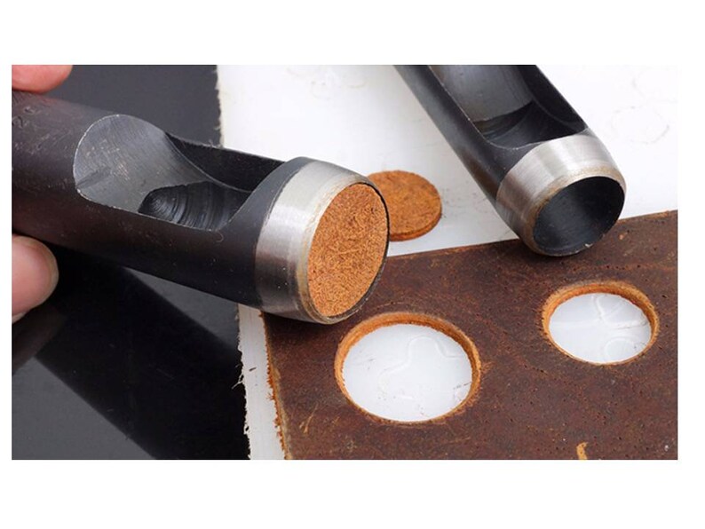 May include: Two black metal leather hole punch tools with a silver rim. The tools are on a brown leather surface with several round holes punched in it. There are also two brown leather circles on the surface.