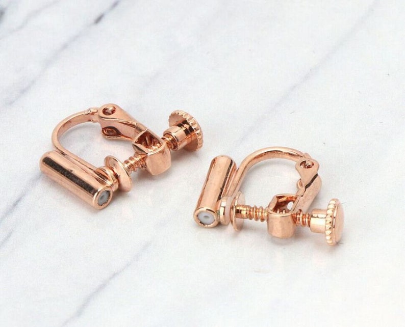 May include: A pair of rose gold clip-on earrings. The earrings have a screw-on back and a cylindrical post.