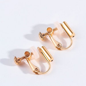May include: A pair of gold-tone clip-on earrings with a screw-on design. The earrings have a white plastic backing.