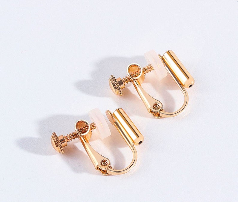 May include: A pair of gold-tone clip-on earrings with a screw-on design. The earrings have a white plastic backing.