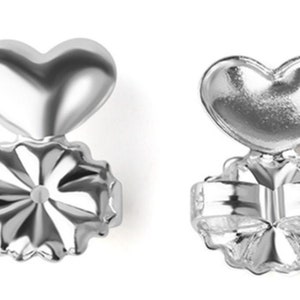 May include: A pair of silver heart-shaped earring backs. The backs are designed to be worn on the back of an earring to secure it to the earlobe. The backs have a heart-shaped design with a small hole in the center for the earring post to go through.