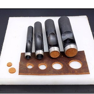 May include: Set of five black leather hole punch tools with different sized punches. The tools are arranged in order of size, with the largest punch on the left and the smallest punch on the right. The punches are on a white surface with a brown leather piece with punched holes.