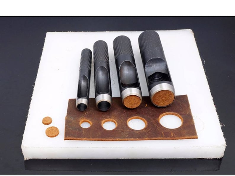 May include: Set of five black leather hole punch tools with different sized punches. The tools are arranged in order of size, with the largest punch on the left and the smallest punch on the right. The punches are on a white surface with a brown leather piece with punched holes.