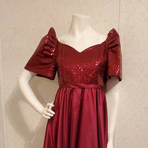 May include: A red sequined bodice with a sweetheart neckline and puff sleeves, attached to a long, flowing red satin skirt. The dress is on a mannequin.