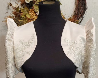 May include: Ivory-colored bolero jacket with short, puffed sleeves. The jacket features intricate floral embroidery along the sleeves and front edges. The bolero is displayed on a black mannequin, showcasing its design and details.