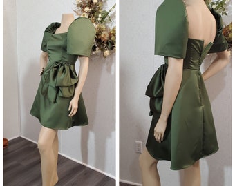 May include: A green satin dress with puffed sleeves and a bow at the waist. The dress has a fitted bodice and a flared skirt. More colors are available.