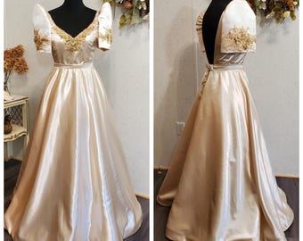 May include: A long, flowing gold satin gown with a V-neckline and short puffed sleeves. The gown features delicate gold embroidery around the neckline and on the sleeves.