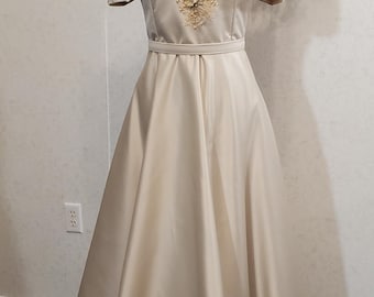 May include: A cream-coloured formal gown with a V-neckline and short sleeves. The bodice features intricate gold embroidery, and the dress has a flowing skirt. The gown is displayed on a black mannequin against a neutral background.