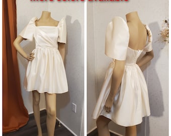 May include: A white satin dress with puffed sleeves and a square neckline. The dress has a fitted bodice and a full skirt. "More colors available" is written in red text at the top of the image.