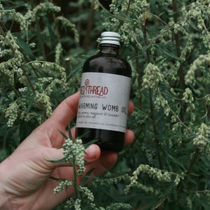 May include: A dark glass bottle with a silver lid labeled "Red Thread" and "Warming Womb Oil" with a list of ingredients including "mugwort & lavender" and "almond oil". The bottle is held in a hand against a backdrop of green foliage.