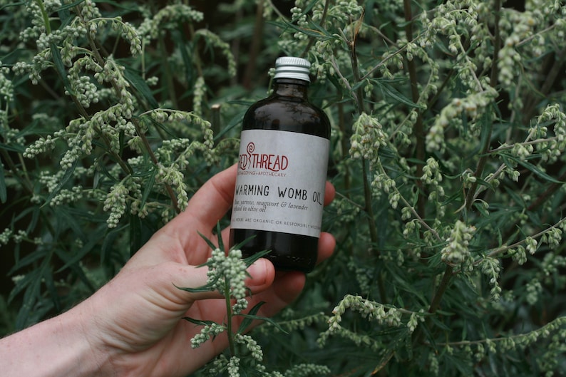 May include: A dark glass bottle with a silver lid labeled "Red Thread" and "Warming Womb Oil" with a list of ingredients including "mugwort & lavender" and "almond oil". The bottle is held in a hand against a backdrop of green foliage.