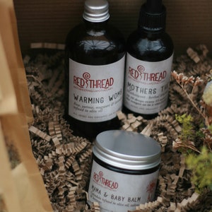 May include: Three glass bottles with silver lids and labels. The bottles contain natural remedies for mothers and babies. The labels read "Red Thread Birthkeeping Apothecary" and include the product names "Warming Womb", "Mothers Tincture", and "Mama & Baby Balm". The bottles are sitting on a bed of brown shredded paper.