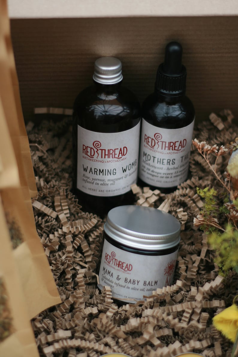 May include: Three glass bottles with silver lids and labels. The bottles contain natural remedies for mothers and babies. The labels read "Red Thread Birthkeeping Apothecary" and include the product names "Warming Womb", "Mothers Tincture", and "Mama & Baby Balm". The bottles are sitting on a bed of brown shredded paper.