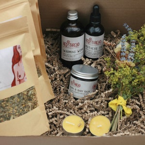 May include: A gift box filled with brown packing paper and a bouquet of dried flowers. The box contains two bags of tea labeled "Milky Mama Breastfeeding Tea", two candles, a bottle of "Warming Woods" oil, a bottle of "Mothers Tincture" oil, and a jar of "Mama & Baby Balm".
