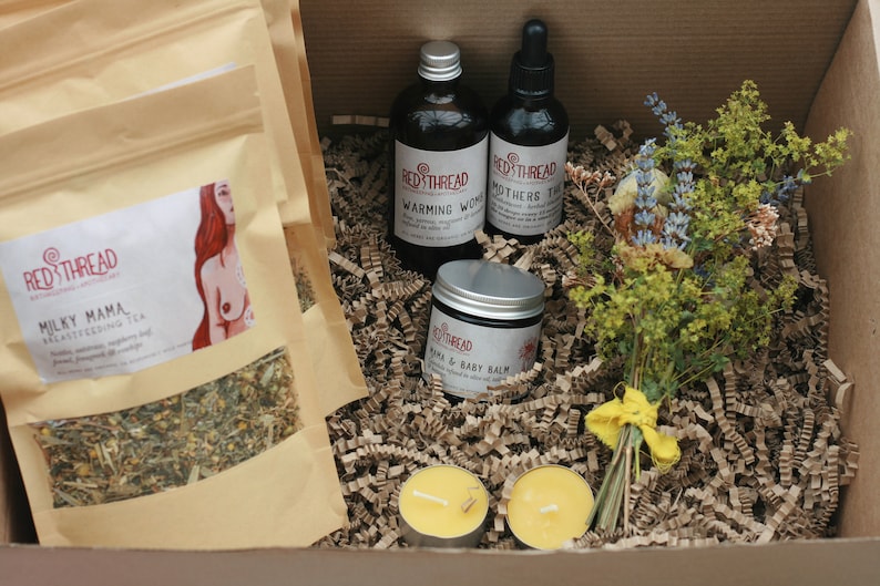 May include: A gift box filled with brown packing paper and a bouquet of dried flowers. The box contains two bags of tea labeled "Milky Mama Breastfeeding Tea", two candles, a bottle of "Warming Woods" oil, a bottle of "Mothers Tincture" oil, and a jar of "Mama & Baby Balm".