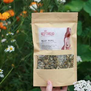 May include: A brown paper bag with a clear window showing a blend of dried herbs. The bag is labeled "Milky Mama Breastfeeding Tea" with a red thread logo and an illustration of a woman breastfeeding.