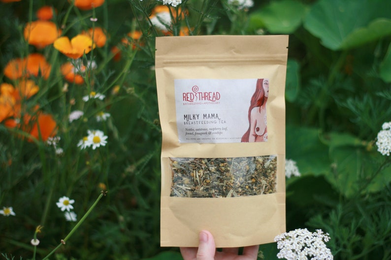 May include: A brown paper bag with a clear window showing a blend of dried herbs. The bag is labeled "Milky Mama Breastfeeding Tea" with a red thread logo and an illustration of a woman breastfeeding.