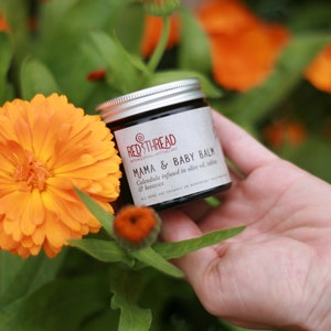 May include: A small jar of Mama & Baby Balm by Red Thread Botanicals. The jar is labeled with the product name and ingredients: Calendula infused in olive oil,  and beeswax. The jar is held in a hand against a background of orange flowers.
