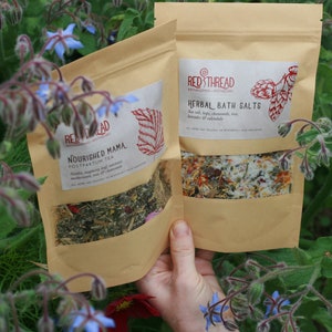 May include: Two brown paper bags with product labels. The bag on the left has the text "Nourished Mama Postpartum Tea" and the bag on the right has the text "Red Thread Herbal Bath Salts".