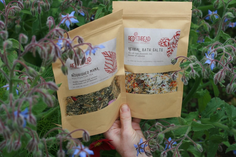 May include: Two brown paper bags with product labels. The bag on the left has the text "Nourished Mama Postpartum Tea" and the bag on the right has the text "Red Thread Herbal Bath Salts".