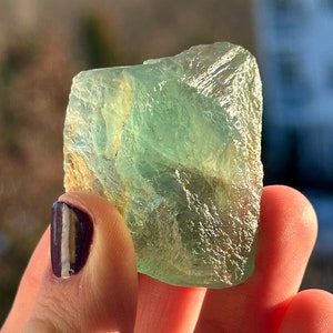 May include: A translucent, light green fluorite crystal with a rough, textured surface. The crystal is held in a hand, with a blurred outdoor background of trees and a building. The crystal's color ranges from pale green to a deeper, more vibrant hue.