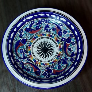 May include: A white ceramic bowl with a blue rim and intricate blue, red, and green floral and fish designs. The bowl has a white center with a black and white starburst pattern.