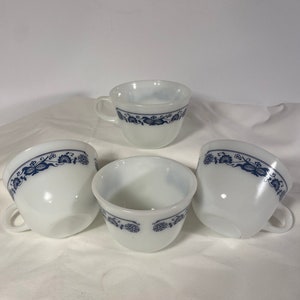 May include: Four white teacups with a blue floral design around the rim. Each cup has a handle and a rounded shape. The cups are arranged on a white surface, showcasing a vintage aesthetic.