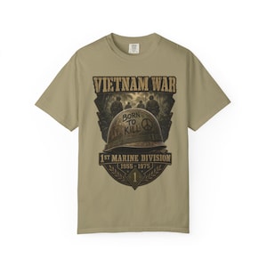 May include: A tan short-sleeve t-shirt featuring a graphic of a military helmet with the text BORN TO KILL and a peace symbol. Above the helmet is VIETNAM WAR and below is 1ST MARINE DIVISION 1955-1975 with a number 1 inside a diamond shape.