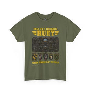 May include: An olive green t-shirt featuring a graphic of a Bell UH-1 Iroquois Huey helicopter instrument panel. The text reads BELL UH-1 IROQUOIS HUEY and WORK HORSES OF VIETNAM, accompanied by five military unit patches in a row below the gauges.