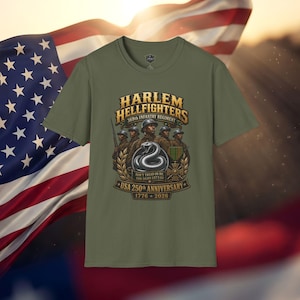 May include: An olive green t-shirt featuring a graphic of the Harlem Hellfighters 369th Infantry Regiment. The design includes soldiers, a coiled snake, a military medal, and the text USA 250th Anniversary 1776-2026, Don't Tread On Me, God Damn Let's Go.