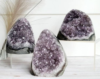 Peut inclure: Trois amas de cristaux d'améthyste, chacun avec une taille et une forme différentes, sont exposés sur une surface blanche. Les cristaux sont d'une couleur violette profonde et ont une texture naturelle et rugueuse.