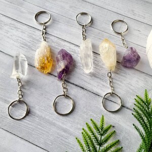 May include: Five crystal keychains with silver rings. The crystals are clear, white, yellow, and purple. The keychains are hanging on a wooden surface.