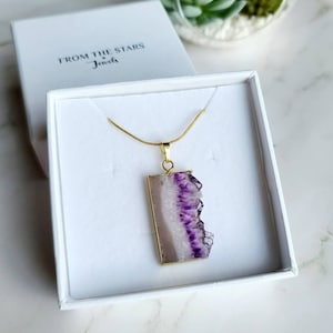 May include: A gold chain necklace with a rectangular pendant featuring a purple and white amethyst stone. The necklace is displayed in a white box with the text "From The Stars Jewels".