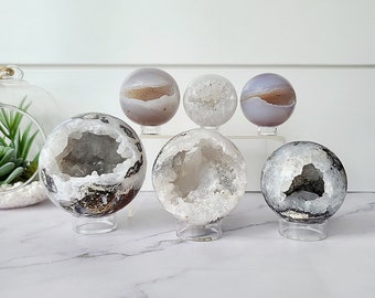 May include: Six agate crystal spheres on clear acrylic stands. The spheres are various sizes and have a natural, white, and gray color with a hollow center.