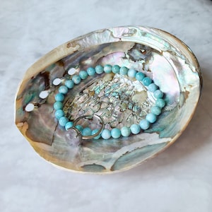 Puede incluir: Un brazalete de piedra de aguamarina azul con un anillo de plata, descansando en una concha de abul&oacute;n natural. La concha tiene un acabado iridiscente y nacarado.