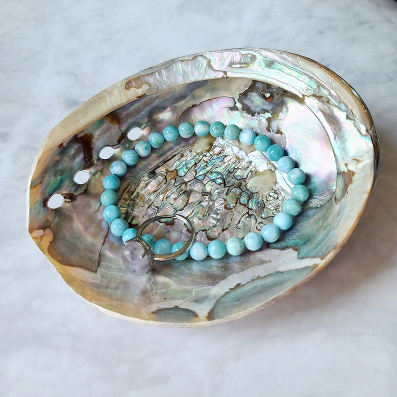 Puede incluir: Un brazalete de piedra de aguamarina azul con un anillo de plata, descansando en una concha de abul&oacute;n natural. La concha tiene un acabado iridiscente y nacarado.