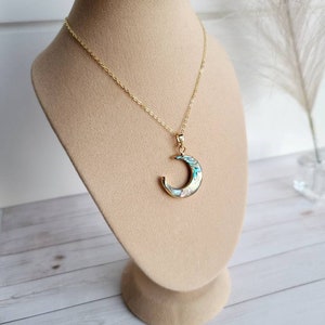 May include: A gold-coloured necklace with a crescent moon pendant. The moon pendant features iridescent abalone shell inlays with blue, white, and green hues. The necklace is displayed on a beige jewellery bust.