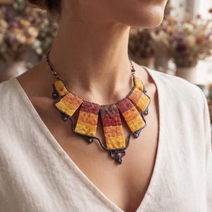 May include: A macrame necklace featuring five rectangular segments in a gradient of yellow, orange, and deep red. The segments are connected by dark brown cord with small metallic beads, forming a curved collar shape around the neck.