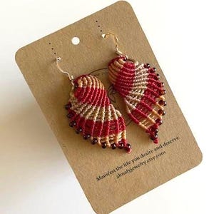 May include: A pair of handmade macrame earrings in a wing design. The earrings feature red and tan woven threads with gold-colored ear wires and small red beads along the bottom edge. The earrings are displayed on a brown card.